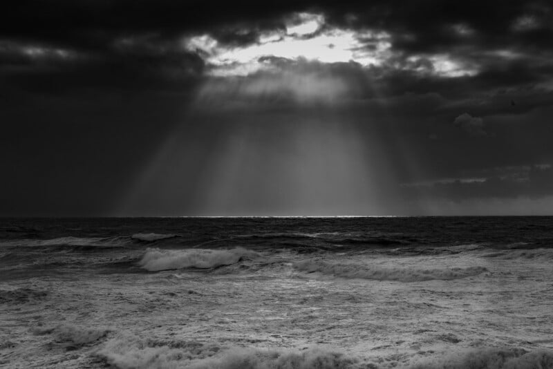 Sunlight breaks through dark, heavy clouds, casting rays down onto a turbulent, choppy ocean in black and white.