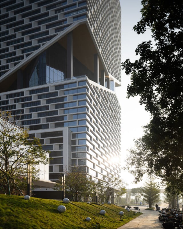 A modern skyscraper with a distinctive geometric facade and a large open-air cutout stands above a grassy slope with decorative spheres and a paved walkway.