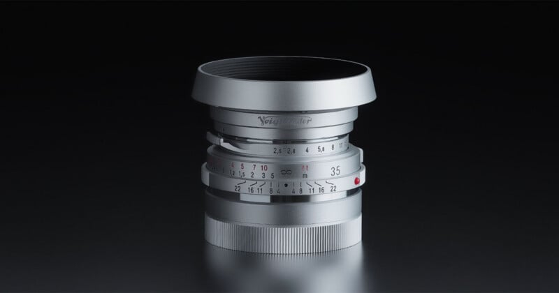 A silver Voigtländer 35mm camera lens with a metal hood stands upright against a dark background.