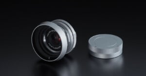A silver Voigtländer Color-Skopar camera lens with a matching lens cap sits on a dark, reflective surface.