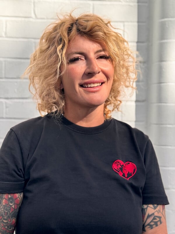 A smiling woman with shoulder-length blonde curly hair wears a black t-shirt featuring a red heart patch with a world map design.