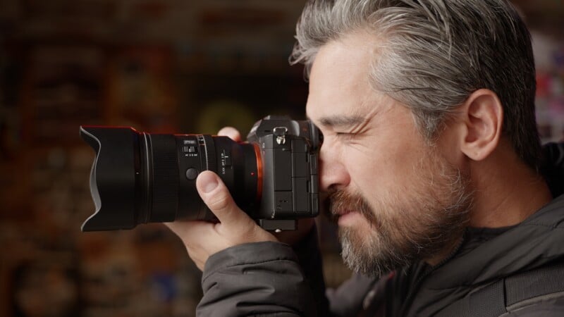 A man with gray hair and a beard holds a camera to his eye, looking through the viewfinder.