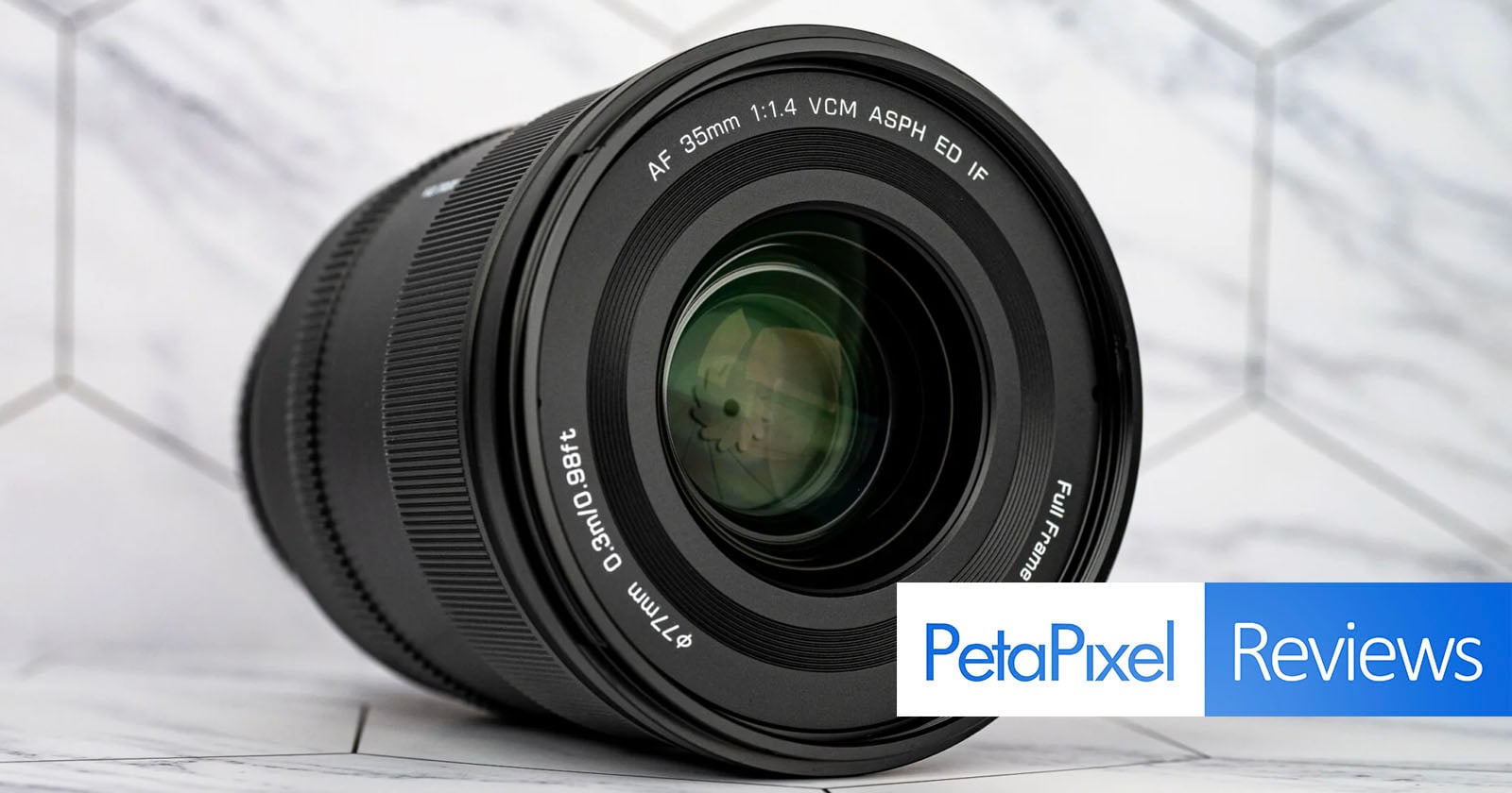 A black camera lens sits on a white marble surface, with the text "AF 35mm 1:1.4 VCM ASPH ED IF" and "Φ77mm 0.3m/0.98ft" visible on the front element.