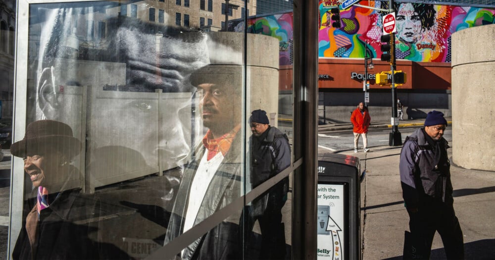 A man in a cap and coat stands behind a glass bus shelter, his reflection overlapping with a large portrait on the glass, while two other men walk on the sidewalk nearby.