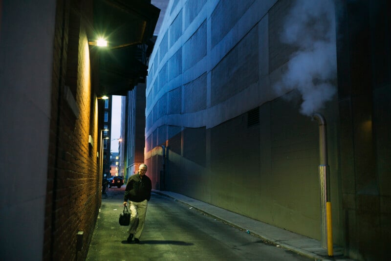An older man carrying a briefcase walks down a narrow, dimly lit urban alleyway past a wall emitting a plume of steam.