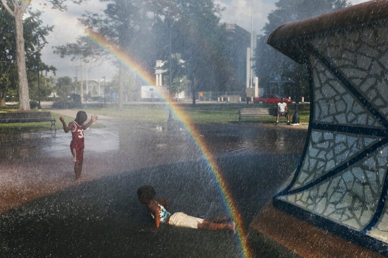 Two children play in the spray of a park fountain, creating a rainbow, while an adult sits on a bench in the background.