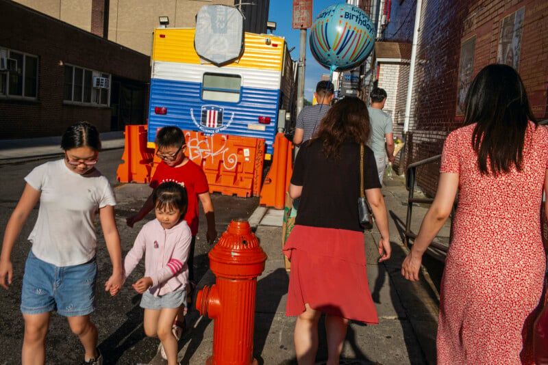 A group of people walk along a sidewalk past a bright orange fire hydrant and a large, blue and yellow trailer parked behind orange construction barriers.
