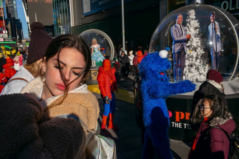 A young woman in a tan fleece jacket stands in a crowded city street near a person in a Cookie Monster costume and a person in a Spider-Man suit.
