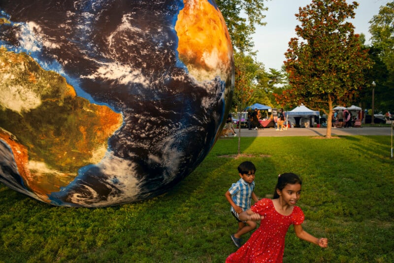 A young girl in a red dress and a boy in a blue plaid shirt run across a grassy park next to a large, inflatable globe.