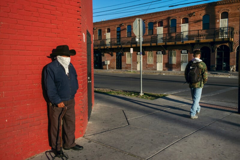 An older man in a cowboy hat and face mask leans against a red brick wall with a cane, while a young man with a backpack walks away on the sidewalk.