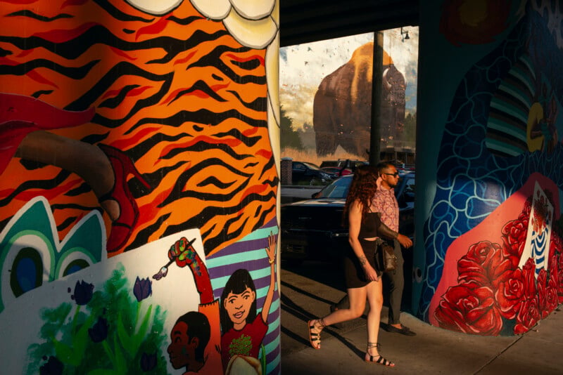 A couple walks past a brightly painted mural pillar, with a large buffalo mural visible in the background.
