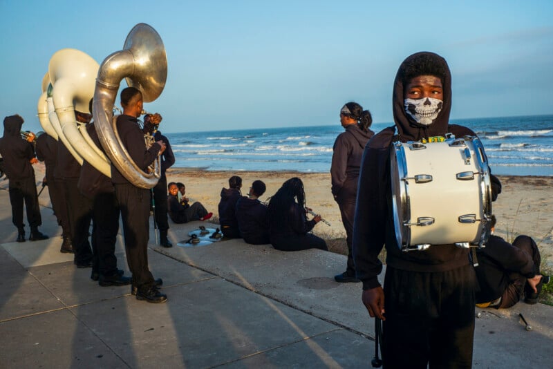 A young person wearing a skull-patterned face mask and carrying a bass drum stands in the foreground, while other band members in black hoodies relax on a concrete path by the ocean.
