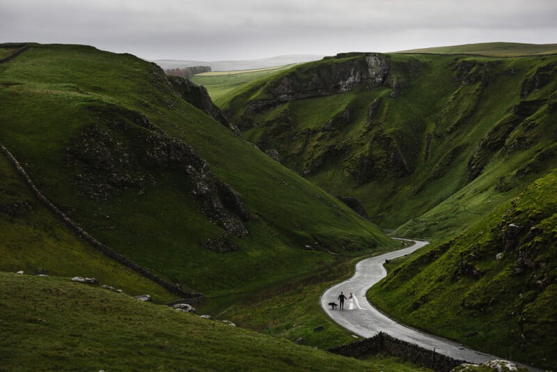 A bride and groom walk hand-in-hand with a dog along a winding road through a lush, green valley under a cloudy sky.