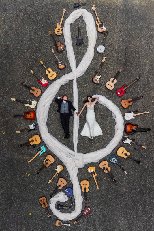 A bride and groom lie on the ground holding hands inside a large treble clef shape formed by white fabric and surrounded by various acoustic and electric guitars.