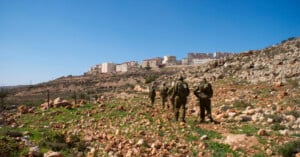 Four soldiers in olive-drab uniforms walk across a rocky, uneven hillside toward a cluster of buildings on the horizon.