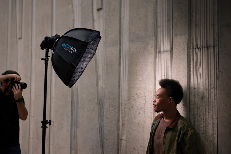 A photographer holds a camera to take a portrait of a man standing against a textured concrete wall, illuminated by a large softbox on a stand.