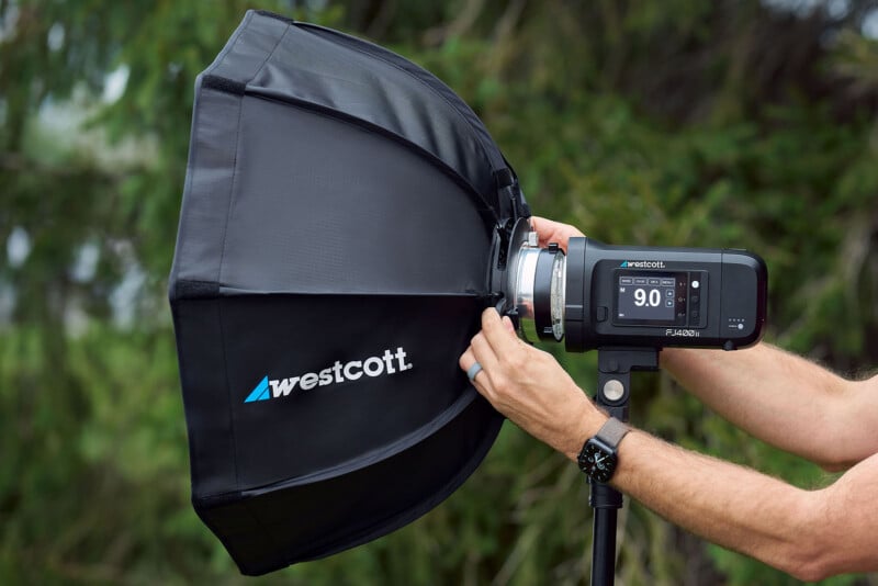 A person's hands adjust a Westcott FJ400 II strobe light with an attached softbox mounted on a light stand outdoors.
