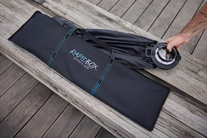 A person's hand pulls a folded RapidBox Switch 4' Octa softbox out of its black carrying case, which is resting on a wooden deck.