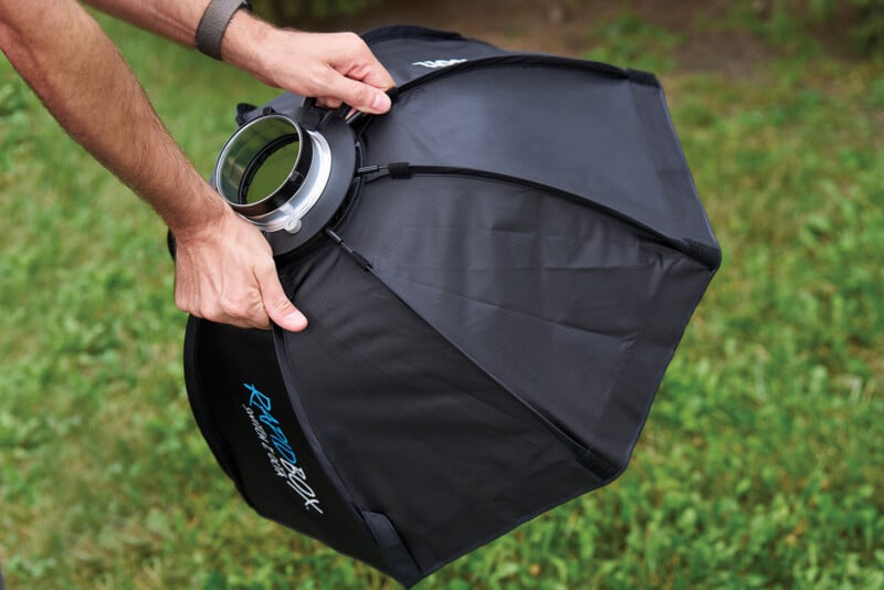 A person holds a black, octagonal photography softbox with a metal mounting ring, set against a background of green grass.