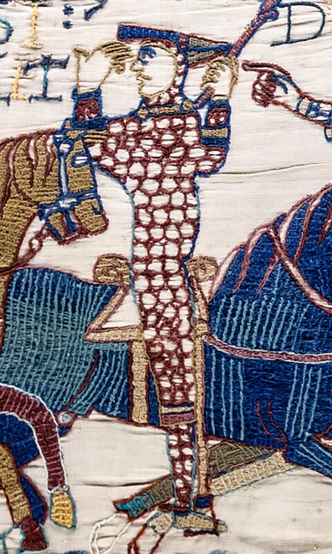 A detail from the Bayeux Tapestry showing a mounted knight in chainmail armor holding a spear, with Latin text visible at the top.