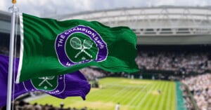Two flags featuring the Wimbledon logo wave in front of a blurred tennis court and stadium crowd.