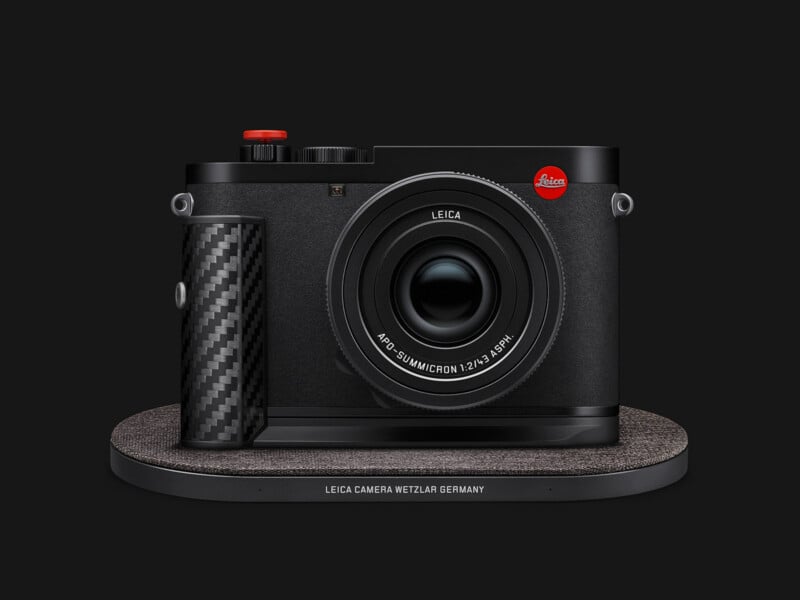 A black Leica camera with a carbon fiber grip and a red shutter button sits on a textured, oval-shaped display base.