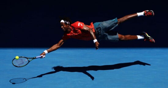 A tennis player in an orange shirt and patterned shorts dives horizontally through the air to hit a yellow tennis ball with his racket.