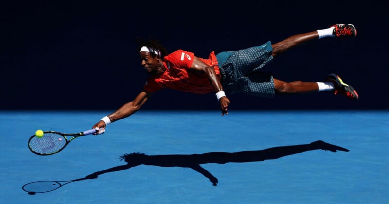 A tennis player in an orange shirt and patterned shorts dives horizontally through the air to hit a yellow tennis ball with his racket.