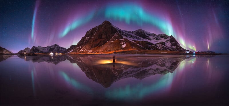 A person stands on a reflective shore at night, silhouetted against a large mountain beneath a vibrant, arched aurora borealis.