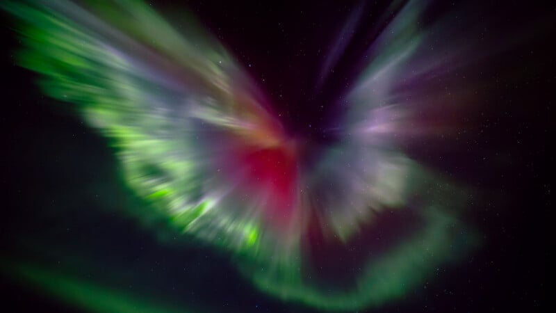 A vibrant aurora borealis displays green, purple, and red light patterns radiating against a dark, star-filled night sky.