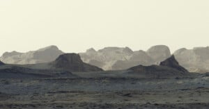 A desolate, rocky landscape under a pale sky, featuring dark, rugged foreground hills and distant, layered mountain ranges.