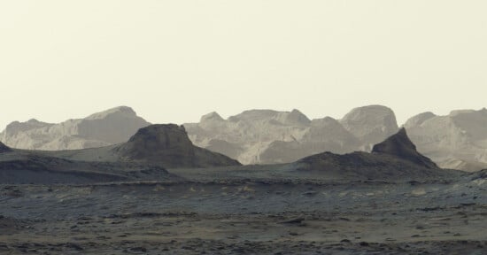 A desolate, rocky landscape under a pale sky, featuring dark, rugged foreground hills and distant, layered mountain ranges.