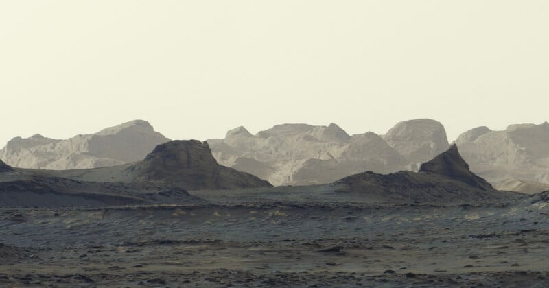 A desolate, rocky landscape under a pale sky, featuring dark, rugged foreground hills and distant, layered mountain ranges.