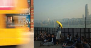 A blurred yellow vehicle passes a brick wall with graffiti, while a group of people sits near a fence overlooking a city.