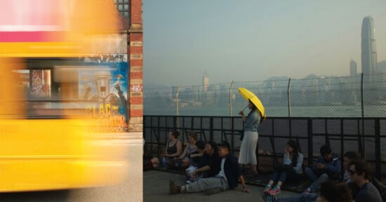 A blurred yellow vehicle passes a brick wall with graffiti, while a group of people sits near a fence overlooking a city.