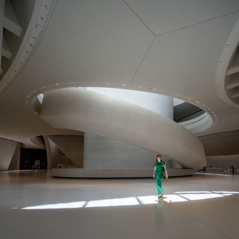 A person in a green outfit walks across a large, minimalist, white-floored hall featuring a prominent curved ramp and circular ceiling cutout.