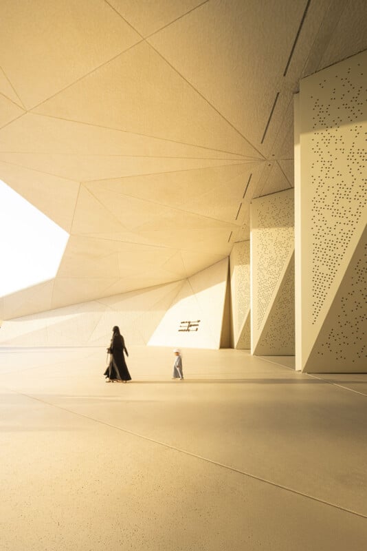 A woman in a black abaya and a young child in traditional clothing walk across a vast, minimalist plaza beneath a large, geometric, light-colored architectural canopy.