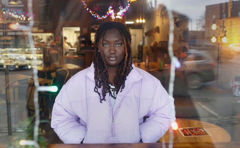 A young person with locs and a nose ring wears a light purple puffer jacket while sitting behind a glass window with fairy lights.