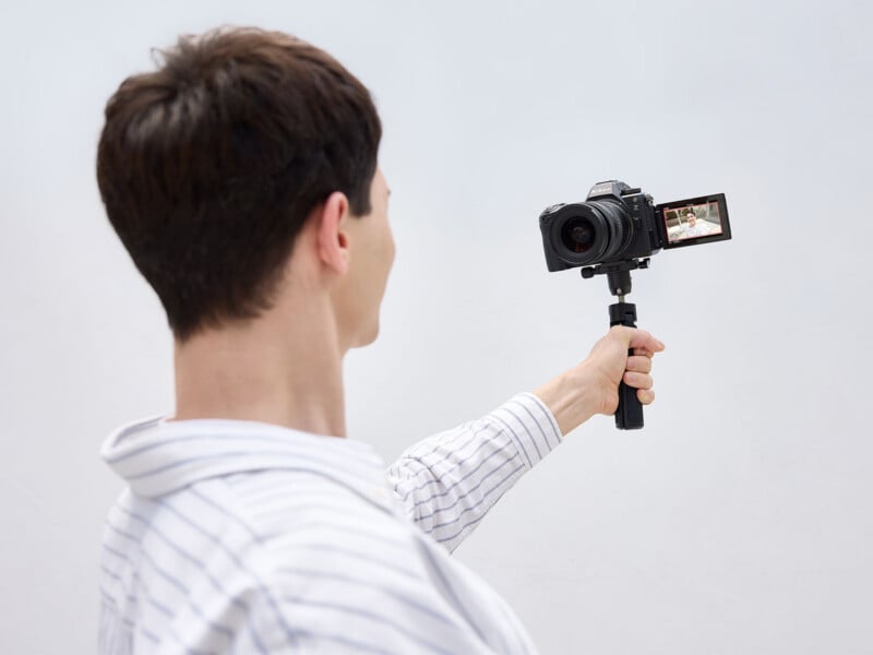 A person seen from behind holds a camera on a small tripod grip, facing the lens toward themselves to view their reflection on the flip-out screen.