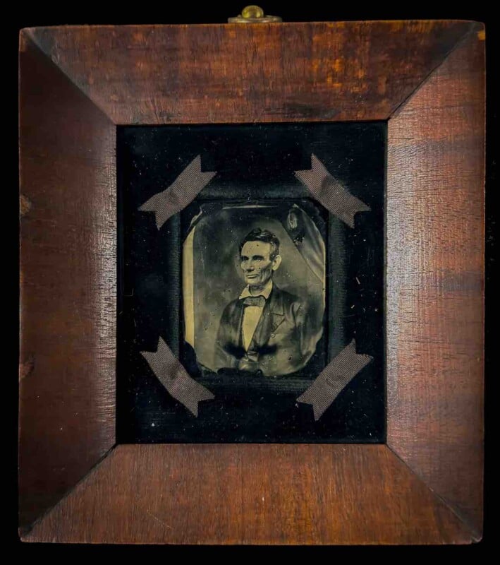 A framed tintype portrait of Abraham Lincoln, mounted on a black background with four decorative ribbons at the corners.