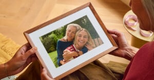 A person holds a framed photograph showing an older woman embracing a younger woman outdoors.