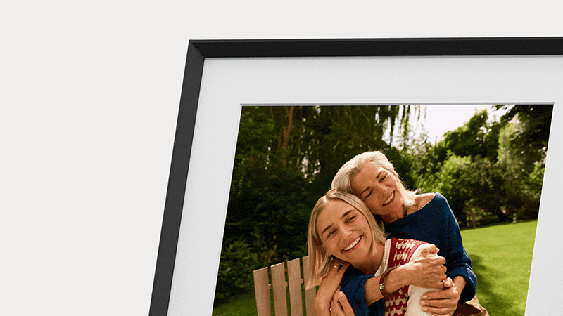 A framed digital display shows an older woman with gray hair hugging a younger woman with blonde hair in a sunny, green backyard.