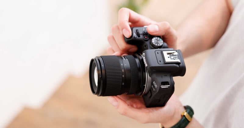 A person holds a black Canon EOS R10 mirrorless camera with a Tamron 18-50mm lens attached.