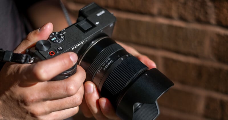A person's hands hold a black Sony mirrorless camera equipped with a Tamron 16-50mm F2.8 lens against a brick wall background.