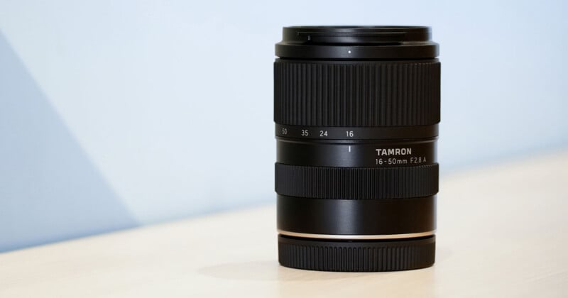 A black Tamron 16-50mm F2.8 camera lens stands upright on a light-colored surface against a plain blue background.