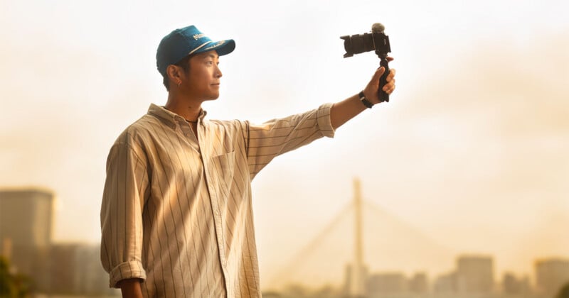 A person wearing a blue baseball cap and a striped button-down shirt holds a camera on a small tripod at arm's length against a hazy, golden-hour cityscape.