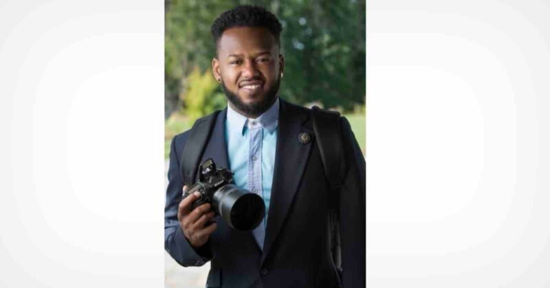 A man with a beard and short dark hair wears a light blue button-down shirt and a dark blazer while holding a camera.
