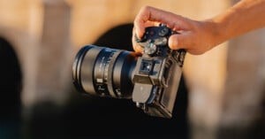 A hand holds a black mirrorless camera with a 35mm f/1.4 lens attached against a blurred, warm-toned background.