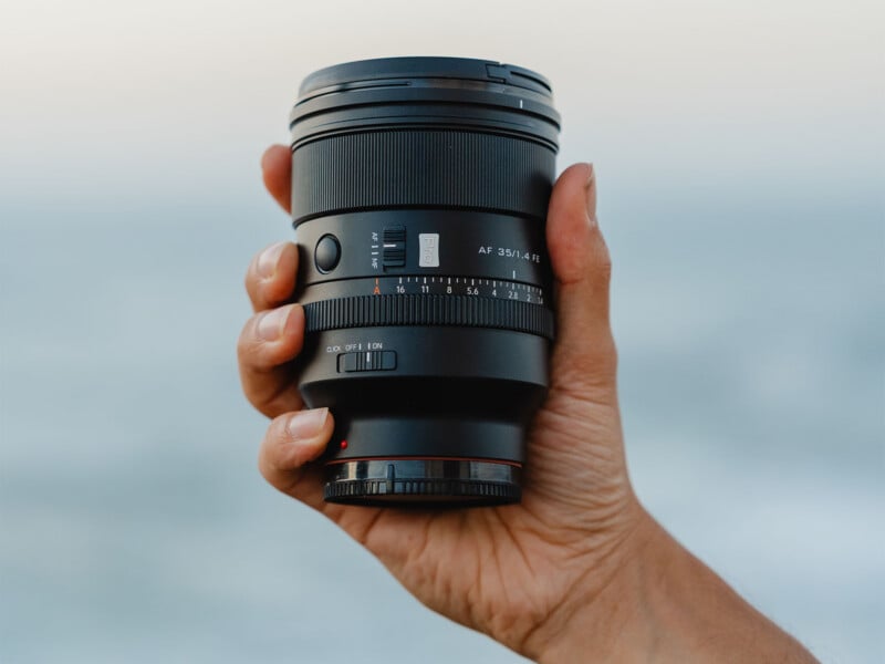 A hand holds a black camera lens against a blurred, light blue background. The lens features an aperture ring, focus switch, and text reading "AF 35/1.4 FE".