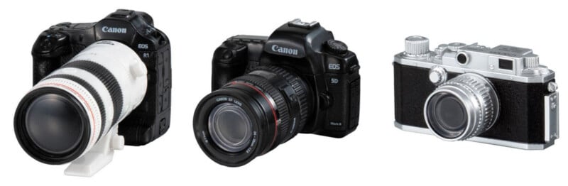 Three miniature Canon cameras are arranged in a row against a white background, featuring a white telephoto lens model, a black DSLR model, and a silver rangefinder-style model.